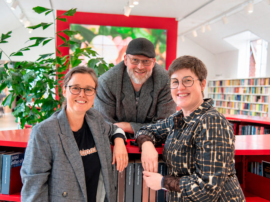 Tre mennesker står samlet i et lyst biblioteksrum.